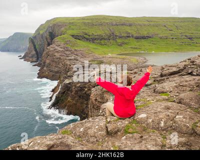 Ein Tourist in roter Jacke sitzt an einem felsigen Ufer über dem Bøsdalafossur Wasserfall, der vom See Sørvágsvatn / Leitisvatn in den Atlantik mündet Stockfoto