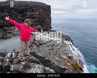 Ein Tourist in roter Jacke sitzt an einem felsigen Ufer über dem Bøsdalafossur Wasserfall, der vom See Sørvágsvatn / Leitisvatn in den Atlantik mündet Stockfoto