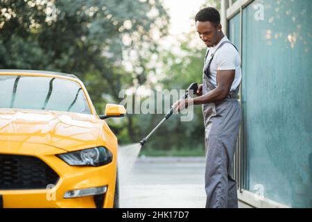 Afrikanischer stilvoller junger Mann mit Fokus auf Arbeitskleidung, der das gelbe Auto mit einer Wasserpistole an der Autostation im Freien wäscht und den Schmutz spült. Stockfoto