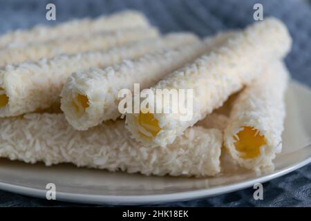 Waffel rollt in Kokosflocken auf einem weißen Porzellanteller. In der Nähe ist ein graues Baumwollküchentuch. Horizontales Foto. Stockfoto