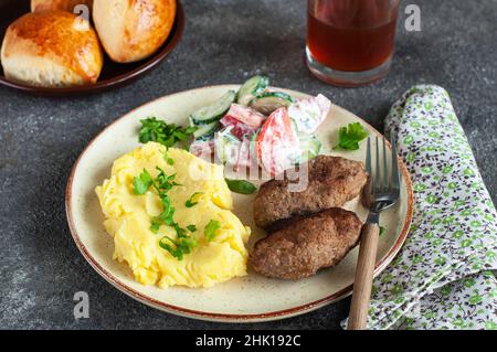 Hausgemachte Fleischschnitzel mit Kartoffelpüree und Gemüsesalat auf einem Teller auf grauem Hintergrund. Stockfoto