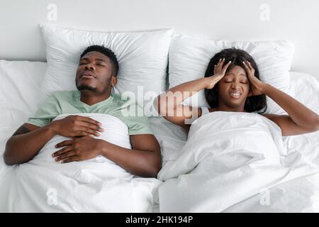 Traurige verzweifelte junge afroamerikanische Frau, die an Kopfschmerzen, Lärm und Schnarchen litt, schlafender Mann im Bett Stockfoto