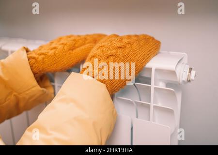 Frauenhänden in gelben gestrickten Fäustlingen berühren einen weißen Heizkörper im Haus. Temperaturprüfung. Stockfoto