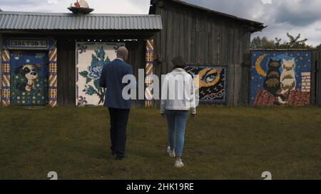 Rückansicht eines Mannes in dunkelblauem Anzug und einer Frau in weißer Jacke und blauer Jeans, der an Holzhäusern mit Mosaikwänden entlang geht. Wände mit wunderschönen MOS Stockfoto