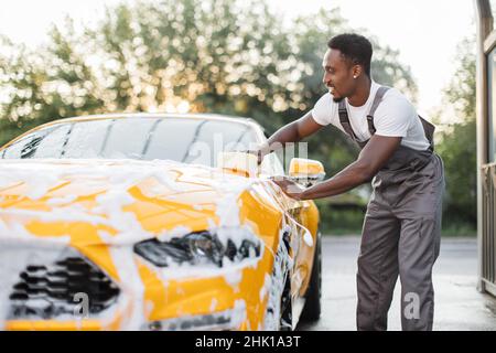 Horizontale Ansicht der Autoreinigung bei der Selbstwäsche im Freien. Hübscher afroamerikanischer Mann in grauen Overalls, der die Motorhaube mit gelbem Schwamm und Schaum wäscht. Stockfoto