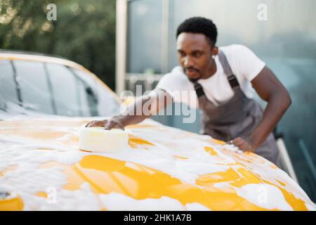 Horizontale Ansicht der Autoreinigung beim Selbstwaschservice. Schöner afrikanischer Mann in T-Shirt und Overalls, der mit einem glücklichen Lächeln auf die Kamera blickt, während er das Auto mit Schwamm und Schaum wäscht Stockfoto