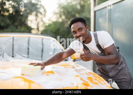 Horizontale Ansicht der Autoreinigung beim Selbstwaschservice. Ein hübscher afrikanischer Mann in T-Shirt und Overalls, der die Kamera anschaut und mit einem glücklichen Lächeln den Daumen nach oben zeigt, während er das Auto mit Schwamm und Schaum wäscht Stockfoto