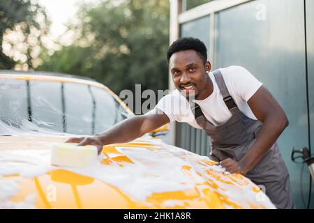 Horizontale Ansicht der Autoreinigung beim Selbstwaschservice. Schöner afrikanischer Mann in T-Shirt und Overalls, der mit einem glücklichen Lächeln auf die Kamera blickt, während er das Auto mit Schwamm und Schaum wäscht Stockfoto