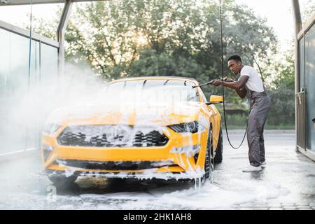 Afrikanischer stilvoller junger Mann mit Fokus auf Arbeitskleidung, der seinen modernen Sportwagen mit einer Wasserpistole an der Autostation im Freien wäscht und den Schaum spült. Stockfoto