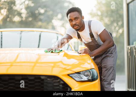 Hübscher junger, beiläufiger afroamerikanischer Mann, der in grauem Overall und weißem T-Shirt die gelbe Autohaube mit grünem Mikrofasertuch bei der Autowäsche unter freiem Himmel abwischt. Stockfoto