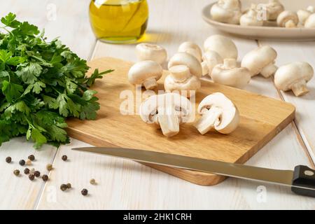Halbierte Rohchampignon- und Küchenmesser auf einem Schneidebrett, Bund Petersilie und ganze Champignons auf einem weißen Holztisch. Pilze kochen. Stockfoto