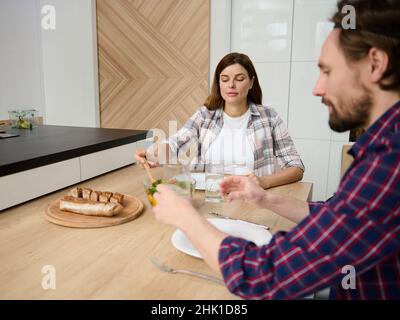 Täglicher Lebensstil eines schönen jungen kaukasischen Paares, das gemeinsam ein Mittagessen genießt und in ihrer geräumigen Villa mit schönem Licht am Tisch sitzt Stockfoto