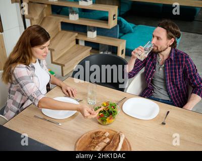 Ein junges kaukasisches Paar isst gemeinsam an einem Tisch zu Mittag. Ein junger Mann trinkt Wasser aus einem Glas, während seine Frau Gemüsesalat auf Teller gießt. Stockfoto