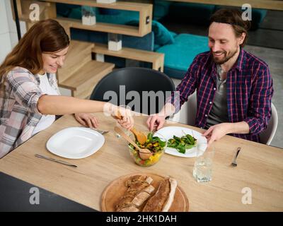 Schöne junge, schwanger Frau, serviert die Frau ihrem Mann einen Gemüsesalat. Glückliches kaukasisches Paar genießen Zweisamkeit beim Mittagessen Stockfoto