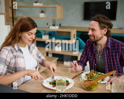 Gutaussehender Mann-Ehemann spricht bei seiner schönen Schwangeren Frau, während er gemeinsam zu Hause leckeres gesundes Mittagessen genießt. Stockfoto