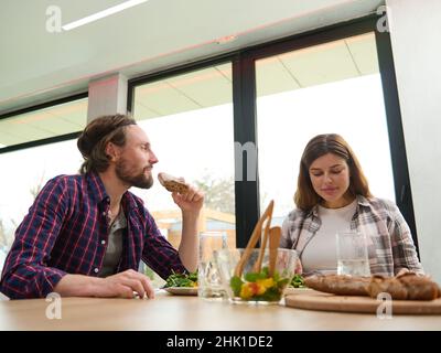 Schöner europäischer Mann und Frau, Mann und Frau, schönes Paar, das sich zu Hause bei einem leckeren Mittagessen am Tisch gegen die Fenster unterhält Stockfoto