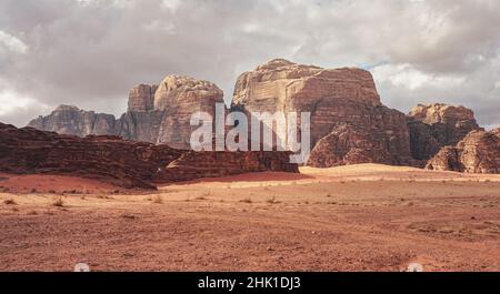 Felsige Massive auf roter Sandwüste, wenig trockener Grashaufen, wolkiger Himmel im Hintergrund, typische Landschaft im Wadi Rum, Jordanien Stockfoto