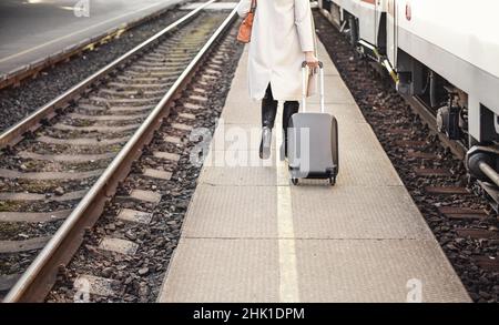 Junge Frau im weißen Mantel, die ihr schwarzes Trolley-Gepäck auf dem Bahnsteig hinter sich zieht, Blick von hinten nur anonymer Unterkörper sichtbar Stockfoto