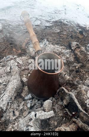 In einer Kupferzezve wird Kaffee auf den heißen Kohlen eines Feuers zubereitet, Dampf kommt von ihm und Schnee liegt um ihn herum. Stockfoto