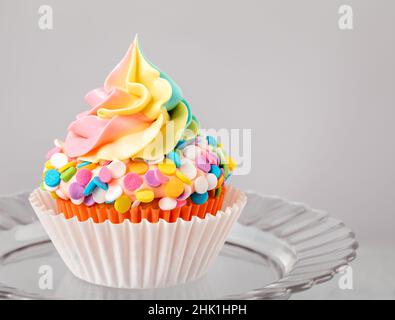 Rainbow Iced Birthday Cupcake auf einem Ständer auf hellgrauem Hintergrund. Stockfoto