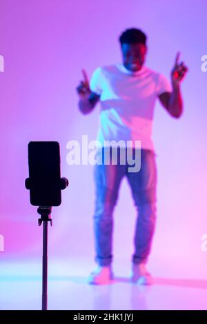 Schwarzer männlicher Influencer, der Video für soziale Medien auf dem Mobiltelefon dreht, Spaß im Neonlicht hat, Nachbau Stockfoto