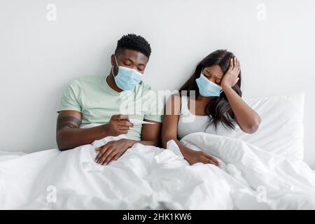 Depressive junge schwarze Dame und Mann in Schutzmasken schauen sich Thermometer auf weißem Bett im Schlafzimmer an Stockfoto