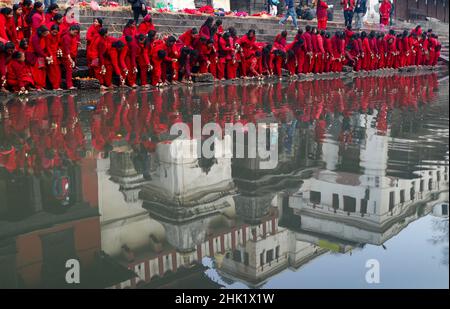 Kathmandu, Nepal. 1st. Februar 2022. Tempelbauten spiegeln sich in den ruhigen Gewässern wider, während sich nepalesische Hindu-Anhänger in roten Gewändern am Ufer des Bagmati-Flusses in der Nähe des Pashupatinath-Tempels während des Madhav Narayan Festivals in Kathmandu versammeln. (Bild: © Aryan Dhimal/ZUMA Press Wire) Stockfoto