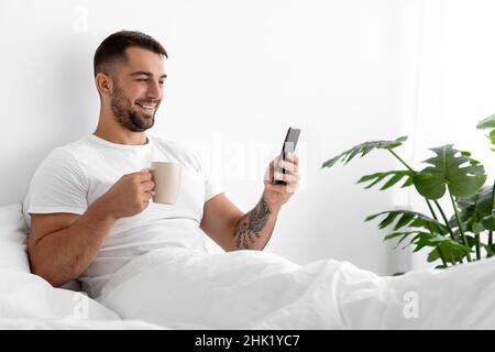 Glücklicher Kaukasisch attraktiver Mann in weißem T-Shirt mit einer Tasse Kaffee im Bett im Schlafzimmer Stockfoto