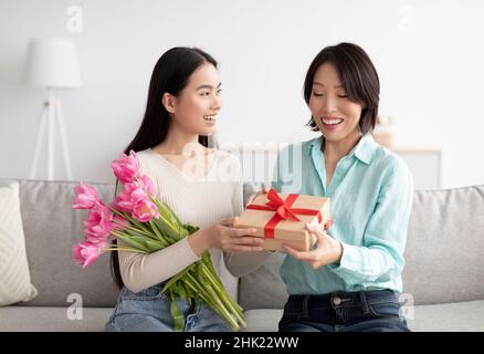 Glückliche Erwachsene asiatische Tochter gibt ihre reife Mutter Blumenstrauß und Geschenk-Box, Gruß sie mit Frauentag zu Hause Stockfoto