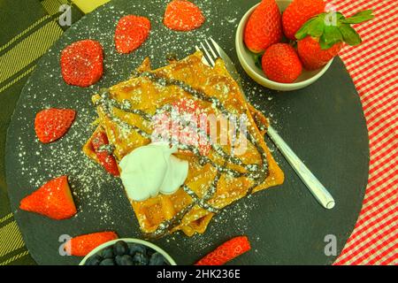 Quadratische belgische Waffeln mit Beeren und Puderzucker auf einem schwarzen Teller. Leckere süße, zuckerhaltige Waffeln. Geschmolzene Schokolade und Schlagsahne auf Waffeln. Oben Stockfoto