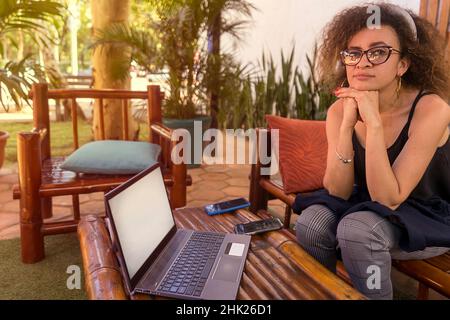 Junge, lockige Latina-Frau mit einem leeren Laptop, die mit einem nervösen Lächeln auf den Lippen auf ihre Geschäfte wartet. Stockfoto
