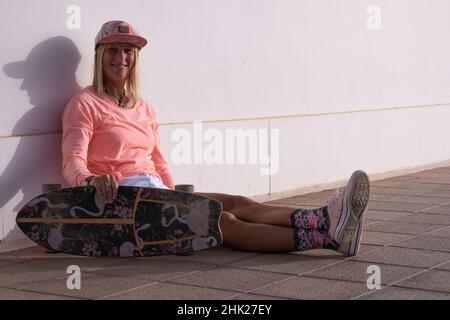 Kaukasische blonde Skaterin mittleren Alters, die auf der Straße sitzt und ihr Skateboard hält und lächelt. Sie trägt eine Hütte. Stockfoto