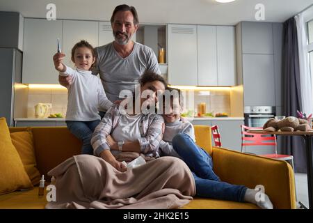 Mutter und Sohn sitzen auf dem Sofa, während Tochter Thermometer zeigt Stockfoto