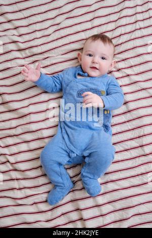 Fröhlicher Junge in blauer Kleidung liegt auf einem gestreiften Bettüberwurf, Draufsicht. Lächelndes Kleinkind im Alter von sechs Monaten im roten Bett, oben Stockfoto