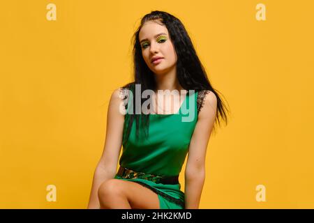 Junge Frau in grünem Kleid sitzt über isolierten gelben Studio-Hintergrund Stockfoto