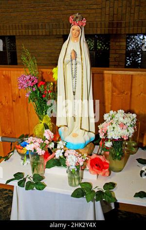 Statue unserer Lieben Frau von Fatima mit Rosenkranz und einer Krone aus Blumen auf ihrer Krone. Sie sitzt auf einem Tisch, umgeben von Schnittblumen Stockfoto