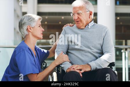 Wie fühlen Sie sich heute? Eine abgeschnittene Aufnahme eines älteren Mannes, der in einem Rollstuhl sitzt, während seine attraktive, reife Krankenschwester ihm in Innenräumen hilft. Stockfoto