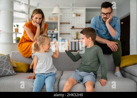 Müde Eltern, die auf der Couch sitzen, fühlen sich erschöpft genervt, während glückliche Kinder zusammen spielen. Stockfoto