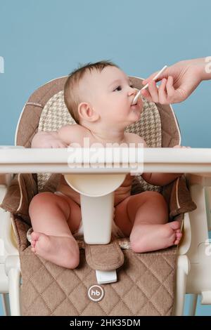 Mutter füttert Kleinkind mit Löffel auf Hochstuhl für die Fütterung von Kindern, blauer Studiohintergrund - Happy Baby, HB - Moskau, Russland, 14. Januar 2022 Stockfoto