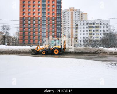 Moskau. Russland. 1. Februar 2022. Ein orangefarbener Traktor mit einem Schaber entfernt Schnee und Vereisung auf einer schmutzigen Straße in der Stadt vor dem Hintergrund mehrstöckiger Gebäude. Stockfoto