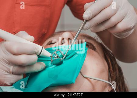 Eine Patientin mit Wangenaufroller mit grüner Kafferdamme in einer Zahnklinik untersucht eine Zahnärztin in Latexhandschuhen ihre Zähne mit einem Bor und einem Spiegel, ein Stockfoto