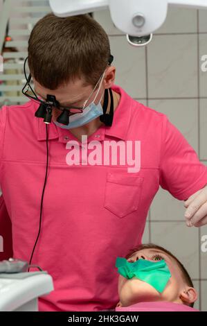 Der Zahnarzt installiert einen Gummidamm und eine Klemme zur Behandlung eines kranken Zahnes, der Junge sitzt in einem Zahnstuhl. Stockfoto