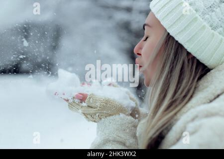 Schönes Mädchen in modischen Winterkleidung bläst Schnee von den Handflächen ihrer Hände. Freude an den ersten Schnee. Junge Blondine in einem weißen Hut in einem verschneiten Stockfoto