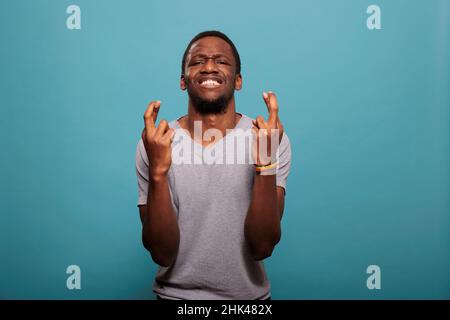 Abergläubische Person, die die Daumen drückt, um um Glück zu beten und auf Erfolg und Sieg zu hoffen. Selbstbewusster Mann, der mit geschlossenen Augen betet, um große Erfolge vor der Kamera zu erzielen. Stockfoto