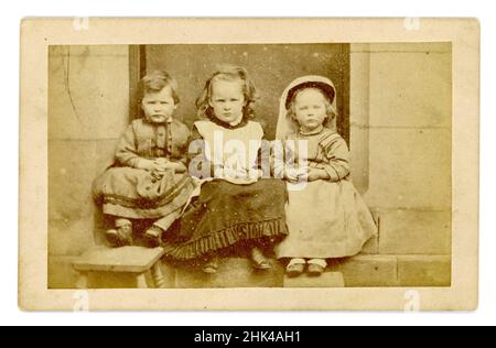 Original viktorianische Carte de Visite (CDV) oder Visitenkarte, von 3 kleinen Kindern, die auf einer Stufe vor einem großen Haus sitzen und jeweils einen Apfel halten. Alle tragen Kleider. Das Kind auf der linken Seite scheint ein Junge zu sein - es war normal, junge Jungen in Mädchenkleidung zu kleiden in viktorianischen Zeiten. Circa 1860, Großbritannien Stockfoto