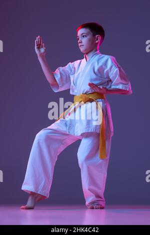 Studio-Aufnahme von sportlichem Kind, männlichem Taekwondo, Karate-Athleten in Doboks, die isoliert auf purpurem Hintergrund in Neon posieren. Konzept des Sports, Kampfkunst Stockfoto