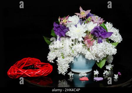 Ein Strauß Frühlingsblumen mit weißem Flieder in einem Keramiktopf und einem roten geflochtenen Herz auf schwarzem Hintergrund mit Reflexen. Valentinstag-Konzept Stockfoto