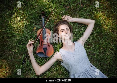 Das Mädchen liegt mit einer Geige auf dem Gras in einem Stadtpark. Stockfoto