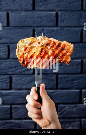 Die Hand hält eine Tranchiergabel mit gegrilltem Schweinekotelett und einer schwarzen Ziegelwand im Hintergrund Stockfoto