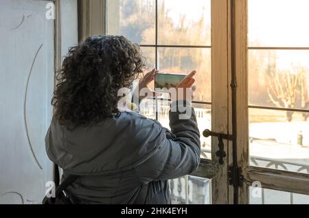 Eine Touristenfrau, die mit ihrem Telefon ein Bild von der Außenlandschaft durch das Glas eines alten Holzfensters eines alten Hauses machte. Stockfoto